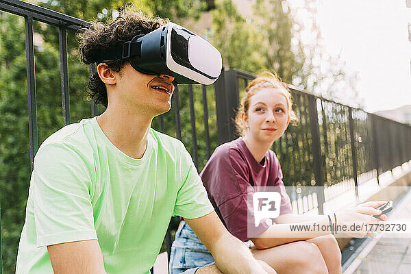 Smiling boy wearing virtual reality simulator sitting by friend in front of railing