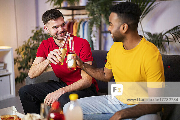 Happy young friends toasting beer bottles sitting at home