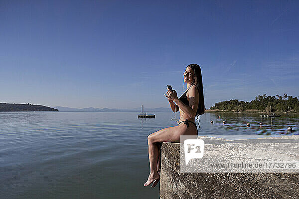 Lächelnde Frau mit Smartphone auf Steg am See