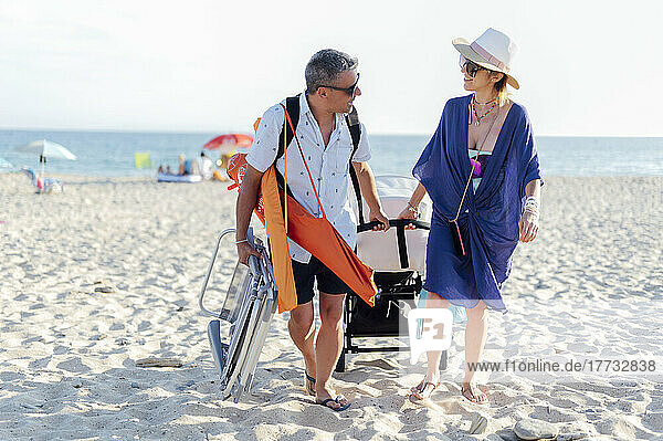 Smiling couple talking with each other pulling baby stroller at beach on sunny day