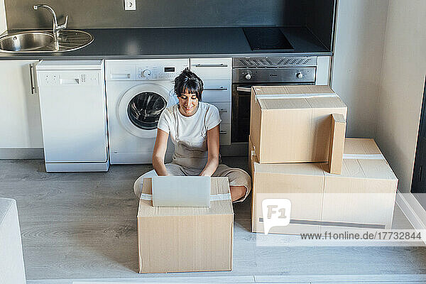 Smiling woman using laptop on cardboard box in front of washing machine at new home