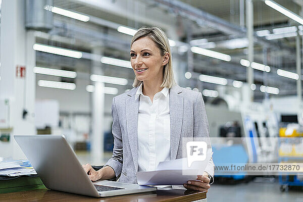 Smiling businesswoman with laptop and documents in factory