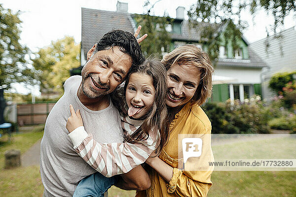Smiling man giving piggyback ride to daughter sticking out tongue by woman in front of house
