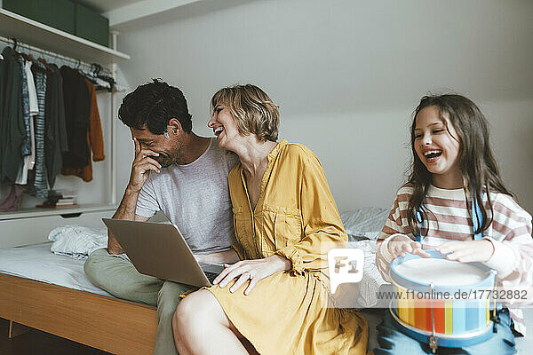 Happy man and woman sitting with laptop by daughter playing toy drum on bed at home