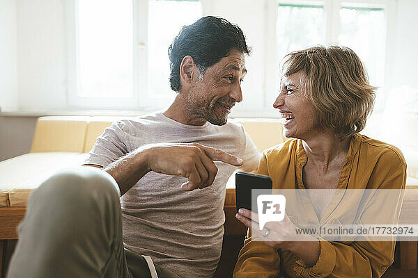 Smiling man talking with cheerful woman holding smart phone at home