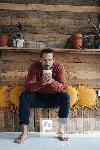 Mann mit geschlossenen Augen hält Tasse in der Hand und sitzt neben Kissen auf Bank