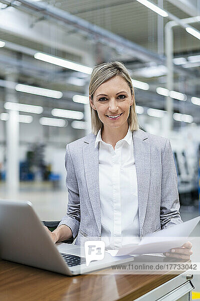 Portrait of smiling businesswoman with laptop and documents in factory