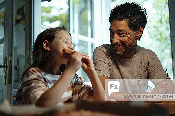 Smiling man looking at daughter eating food at home