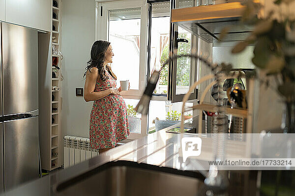 Smiling pregnant woman with tea mug standing in kitchen looking through window