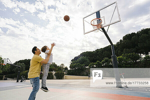 Father carrying son throwing basketball at sports court