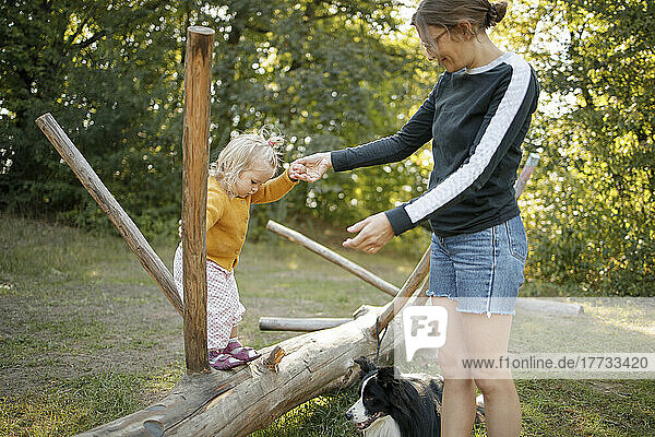 Girl holding hand of mother playing on fallen tree by dog in forest