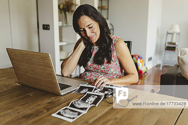 Smiling pregnant woman sitting with laptop looking at ultrasound scans