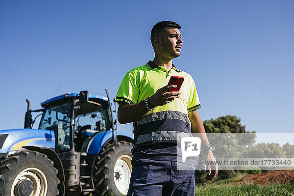 Farmer holding mobile phone in front of tractor