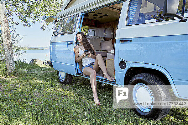 Woman with mobile phone day dreaming in motor home by lake