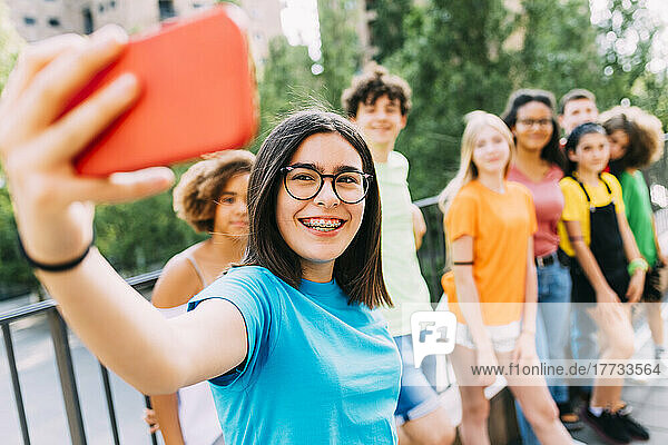 Cheerful girl wearing eyeglasses taking selfie through smart phone with friends