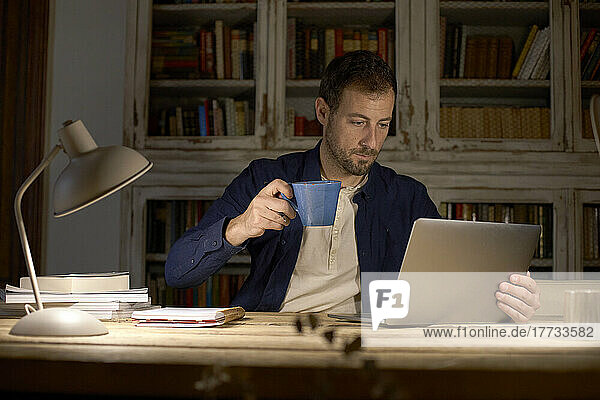 Businessman holding coffee cup working on laptop at home