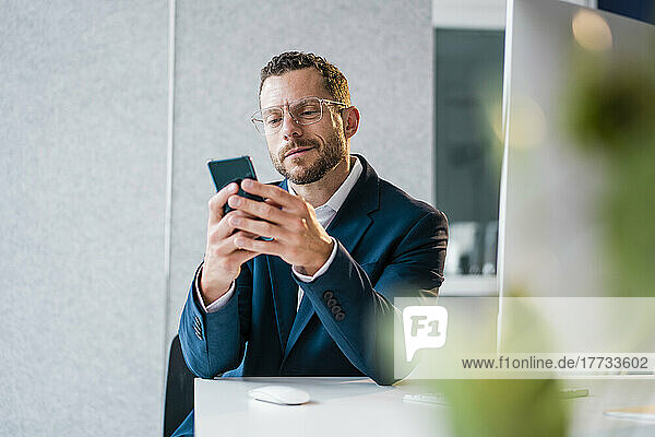 Smiling businessman using smart phone sitting at desk in office