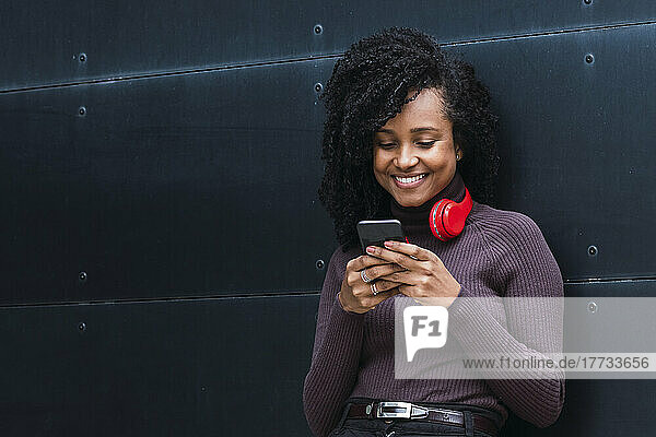 Smiling woman wearing wireless headphones using smart phone in front of wall