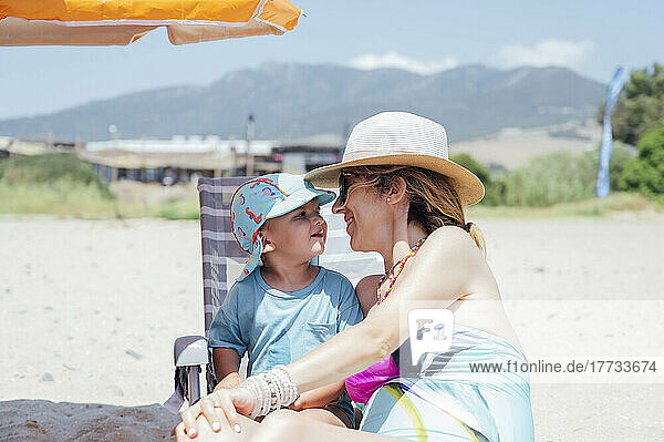 Happy mother wearing hat sitting with son at beach on sunny day