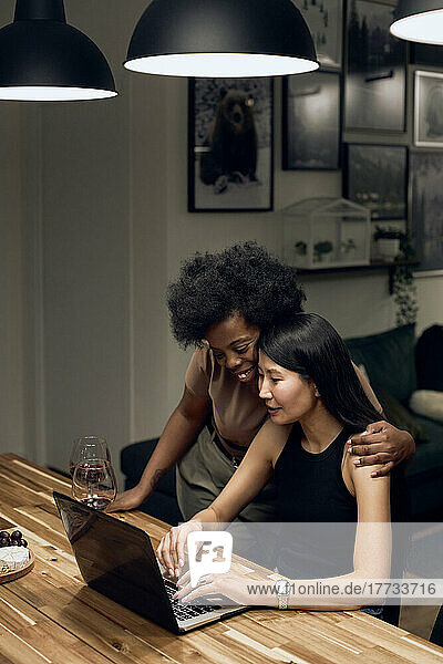Smiling woman embracing girlfriend using laptop sitting at table