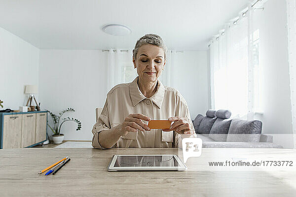 Frau sitzt zu Hause am Tisch  hält Karte in der Hand und nutzt digitales Tablet
