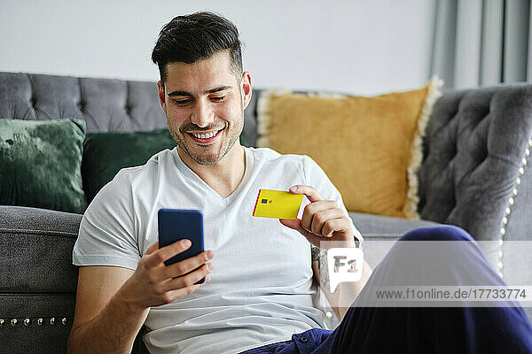Smiling man holding credit card doing online shopping through smart phone at home
