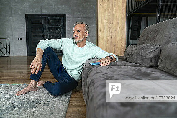 Thoughtful man with mobile phone sitting by sofa at home