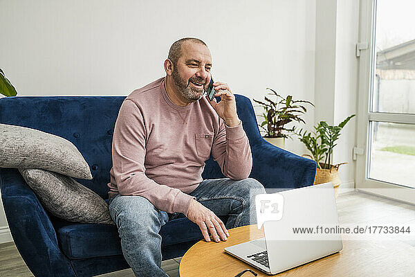 Lächelnder Geschäftsmann  der mit einem Laptop im Wohnzimmer sitzt und am Smartphone spricht