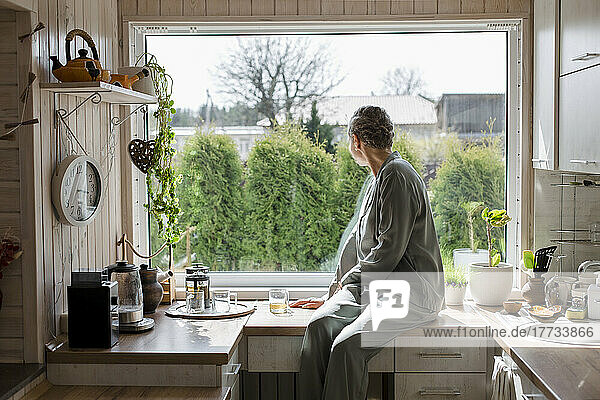 Mature woman sitting on kitchen counter at home looking out of window