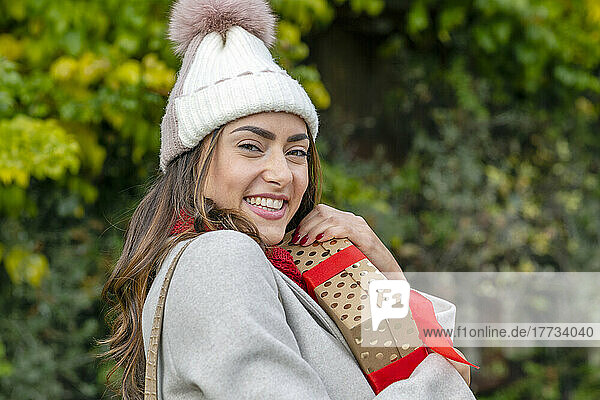 Beautiful happy woman wearing knit hat hugging Christmas present at park