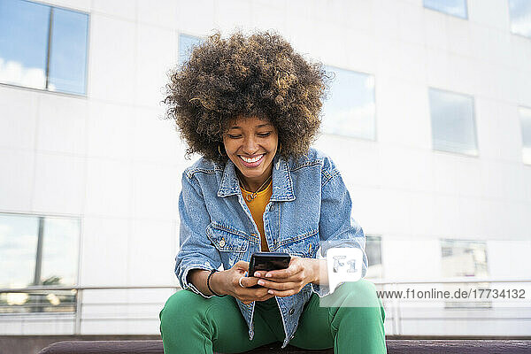 Cheerful Afro woman text messaging through smart phone sitting on bench