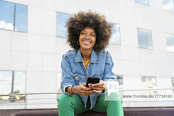 Beautiful happy woman with Afro hairstyle holding mobile phone sitting on bench