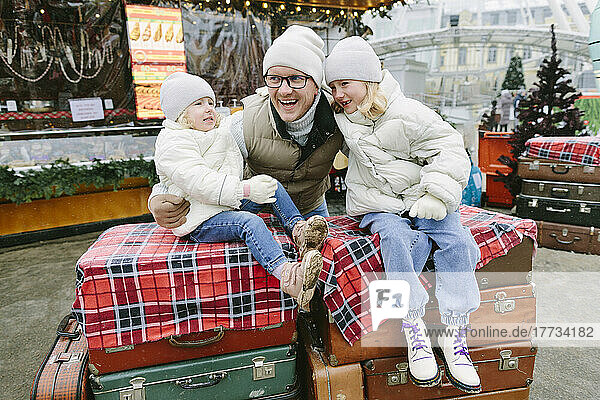 Smiling girls sitting on suitcases by father at Christmas market