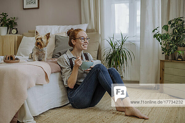 Happy woman with bowl of food sitting by dog on bed at home