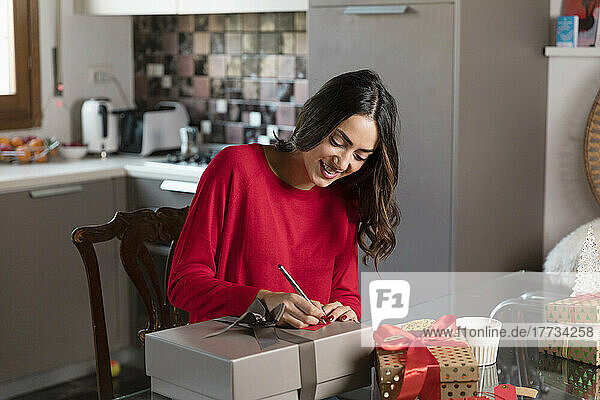 Lächelnde junge Frau schreibt zu Hause auf einen Geschenkanhänger für ein Weihnachtsgeschenk