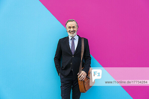 Happy mature businessman wearing bag standing in front of pink and blue wall