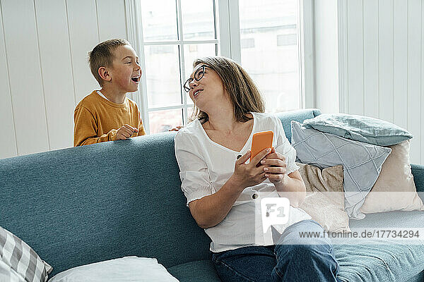 Fröhlicher Junge spricht mit seiner Mutter  die zu Hause auf dem Sofa sitzt und ihr Mobiltelefon in der Hand hält