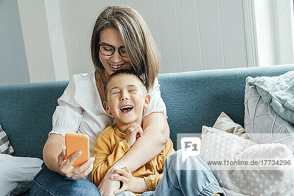 Happy boy with mother holding smart phone sitting on sofa at home