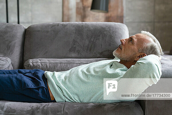 Thoughtful man with hands behind head lying on sofa at home
