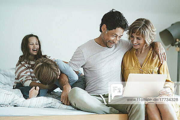 Glücklicher Mann und Frau mit Laptop sitzen neben Tochter und Sohn und spielen zu Hause im Bett