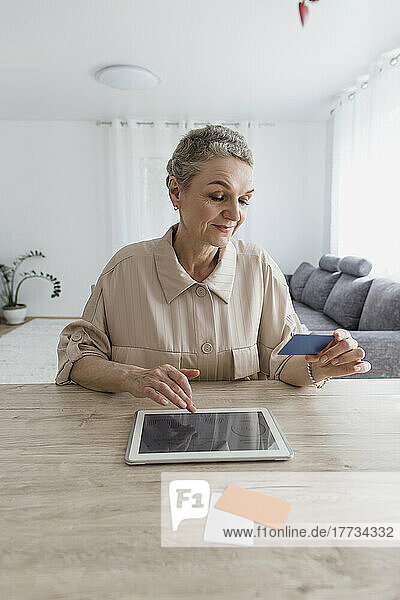 Frau sitzt zu Hause am Tisch  hält Karte in der Hand und nutzt digitales Tablet