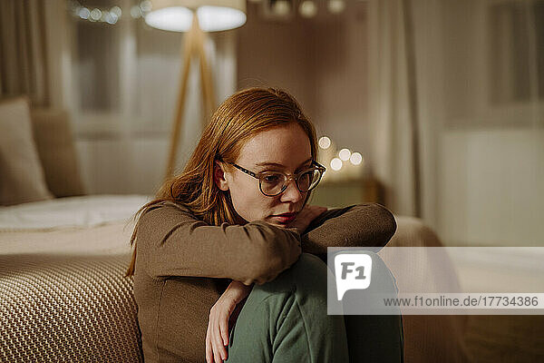 Sad woman wearing eyeglasses sitting in front of bed at home