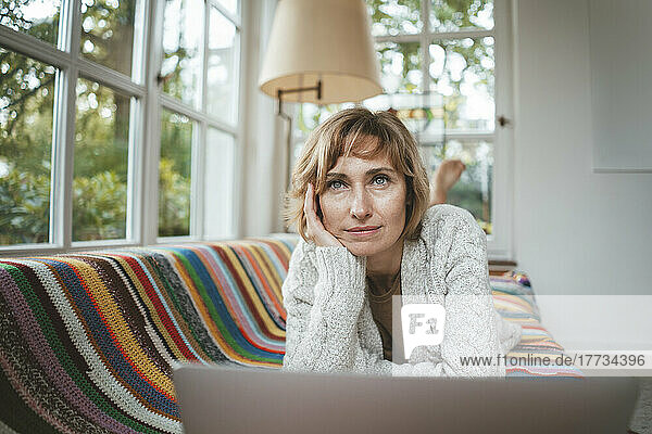 Thoughtful mature woman with laptop lying on sofa at home