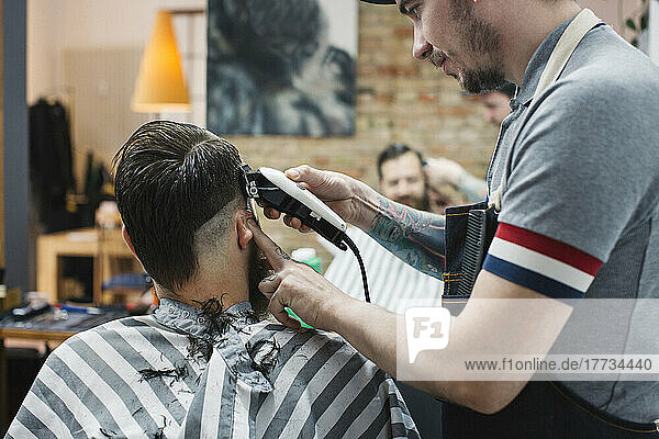 Hairdresser cutting hair of man with electric razor at barber shop