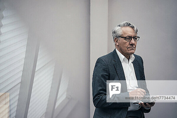 Thoughtful senior businessman with tablet PC standing in front of wall