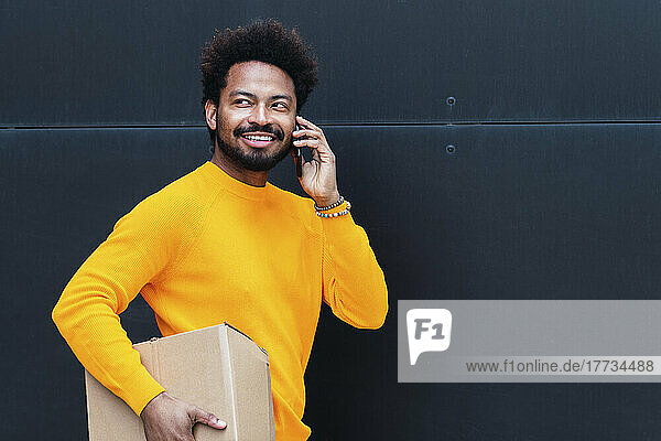 Smiling delivery man holding box talking on smart phone in front of black wall