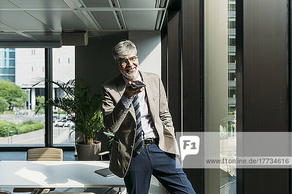 Happy businessman talking on smart phone through speaker standing by window at office