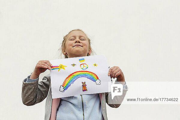 Mädchen mit geschlossenen Augen zeigt Regenbogen und Friedenssymbol auf Papier vor weißem Hintergrund