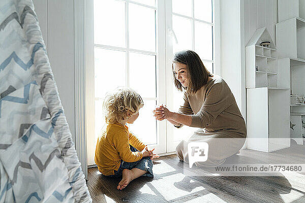 Happy mother playing with son sitting by window at home