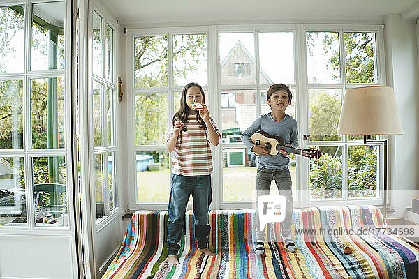 Siblings playing musical instrument standing on sofa at home
